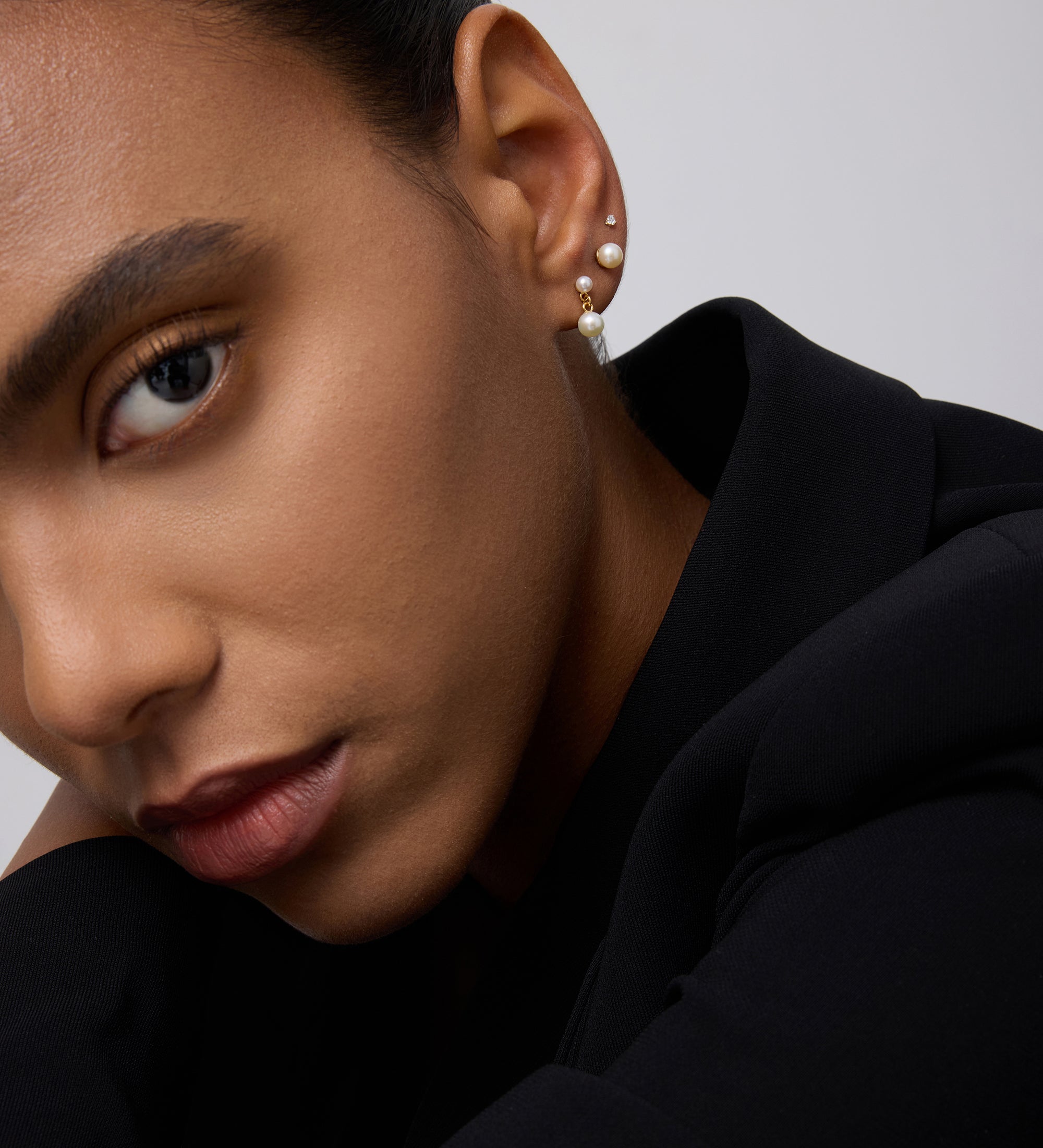 A close-up of a person’s face in a black outfit, highlighting smooth skin, groomed brows, neutral makeup, and three earrings—one is the 14K Solid Yellow Gold Lab-grown Diamond Prong Set Flat Back Stud (2mm)—against a plain light background.