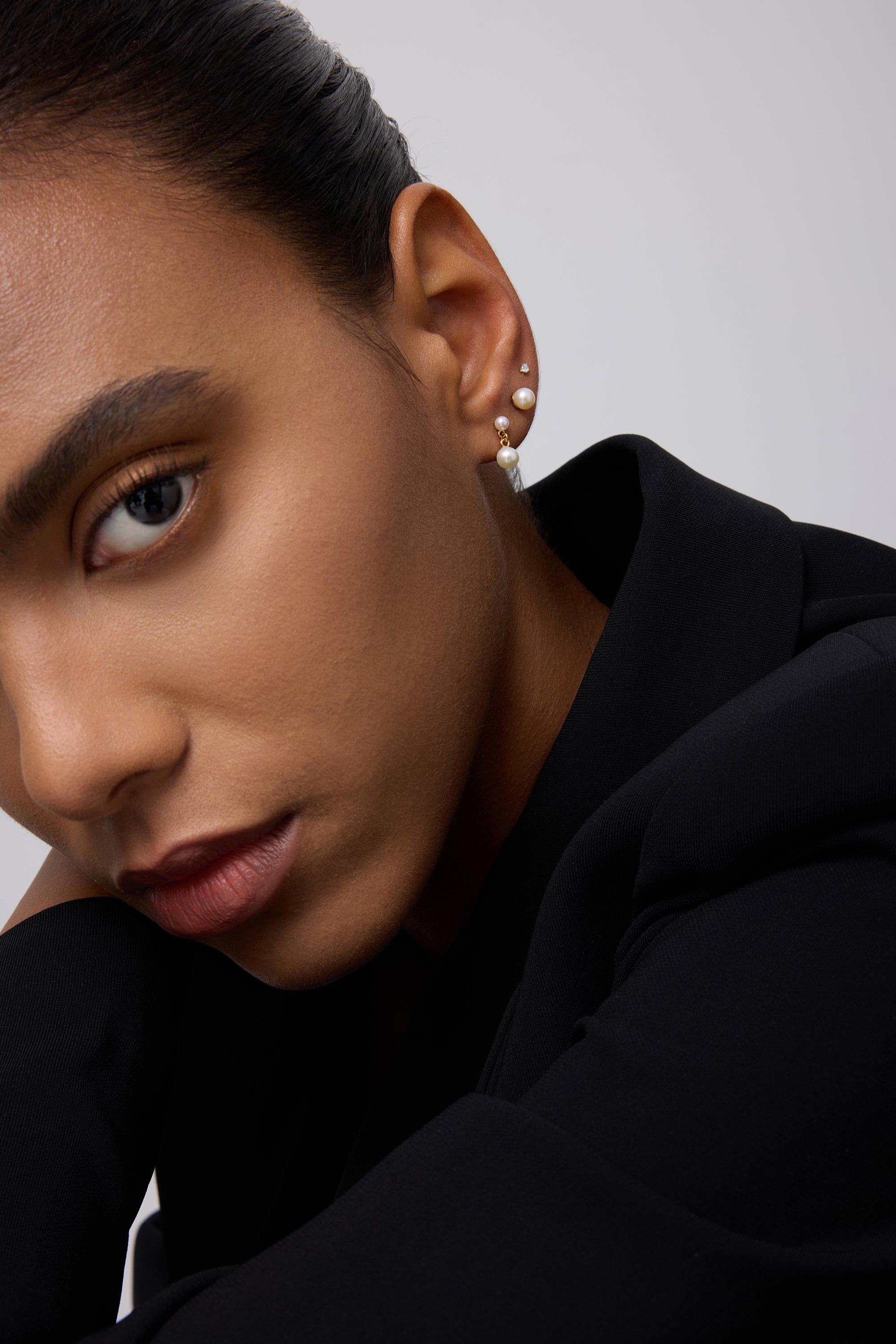 A close-up of a person’s face in a black outfit, highlighting smooth skin, groomed brows, neutral makeup, and three earrings—one is the 14K Solid Yellow Gold Lab-grown Diamond Prong Set Flat Back Stud (2mm)—against a plain light background.
