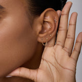 A close-up of a person holding their hand to their ear showcases the 14K Solid Yellow Gold Lab-grown Diamond Double Chain Flat Back Stud among other piercings. The background is neutral, with only part of the face in view.