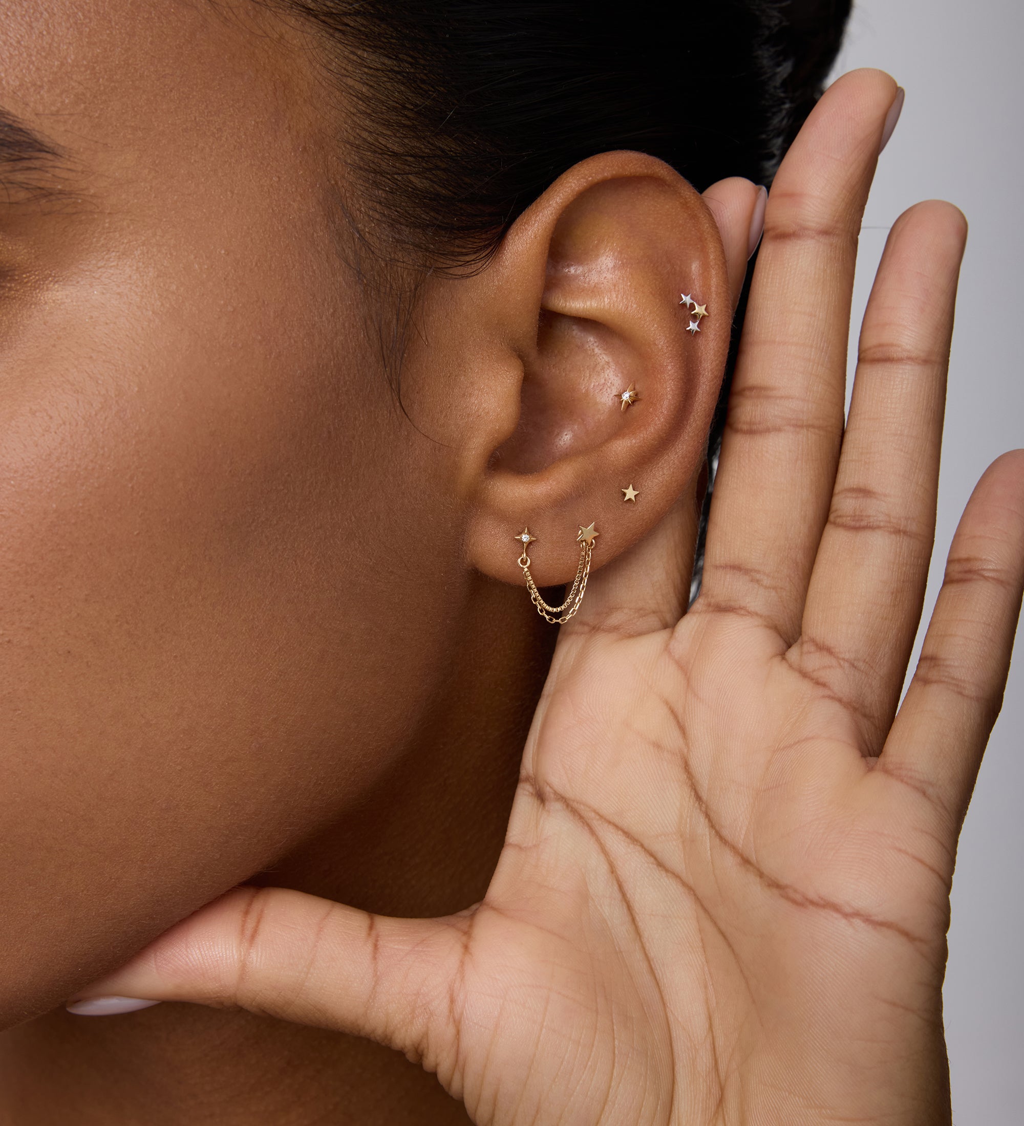 A close-up of a person holding their hand to their ear showcases the 14K Solid Yellow Gold Lab-grown Diamond Double Chain Flat Back Stud among other piercings. The background is neutral, with only part of the face in view.