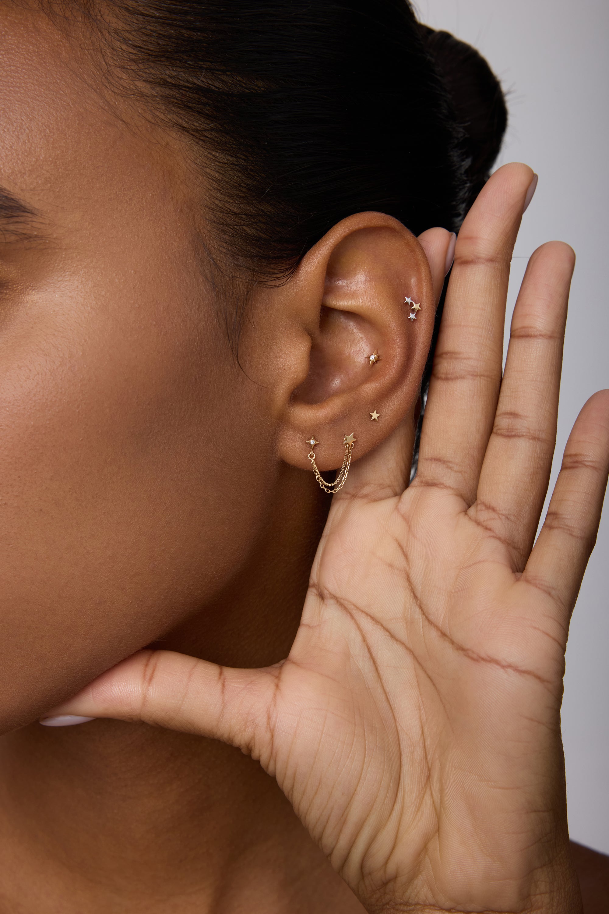 A close-up of a person holding their hand to their ear showcases the 14K Solid Yellow Gold Lab-grown Diamond Double Chain Flat Back Stud among other piercings. The background is neutral, with only part of the face in view.