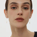 A person with fair skin and dark hair pulled back is wearing subtle makeup and the Eden Trio Pearl Drop Earrings in Silver. They are dressed in a sleeveless black top and looking directly at the camera against a plain background.
