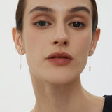 A person with fair skin and dark hair pulled back is wearing subtle makeup and the Eden Trio Pearl Drop Earrings in Silver. They are dressed in a sleeveless black top and looking directly at the camera against a plain background.