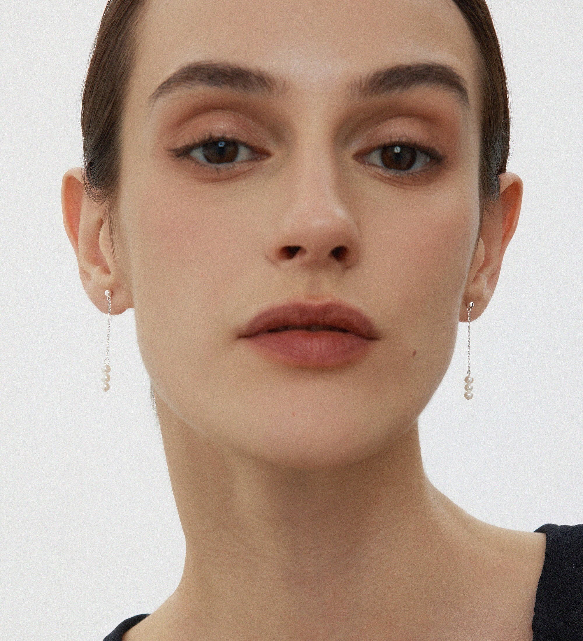 A person with fair skin and dark hair pulled back is wearing subtle makeup and the Eden Trio Pearl Drop Earrings in Silver. They are dressed in a sleeveless black top and looking directly at the camera against a plain background.