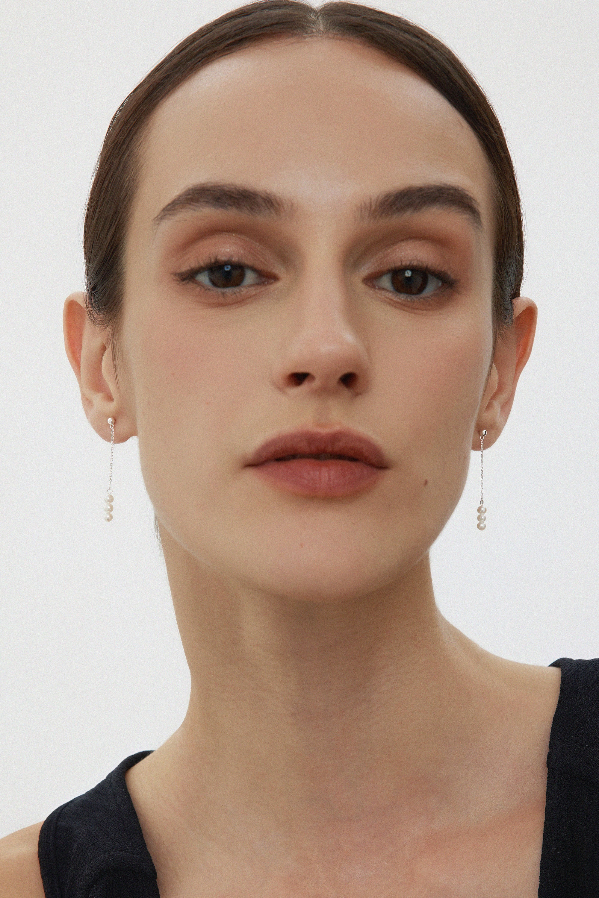 A person with fair skin and dark hair pulled back is wearing subtle makeup and the Eden Trio Pearl Drop Earrings in Silver. They are dressed in a sleeveless black top and looking directly at the camera against a plain background.