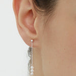 Close-up of a womans ear wearing the Eden Trio Pearl Drop Earrings in Silver, featuring a small stud and three white pearls dangling vertically from a thin silver chain. These elegant earrings stand out against a plain, neutral background.