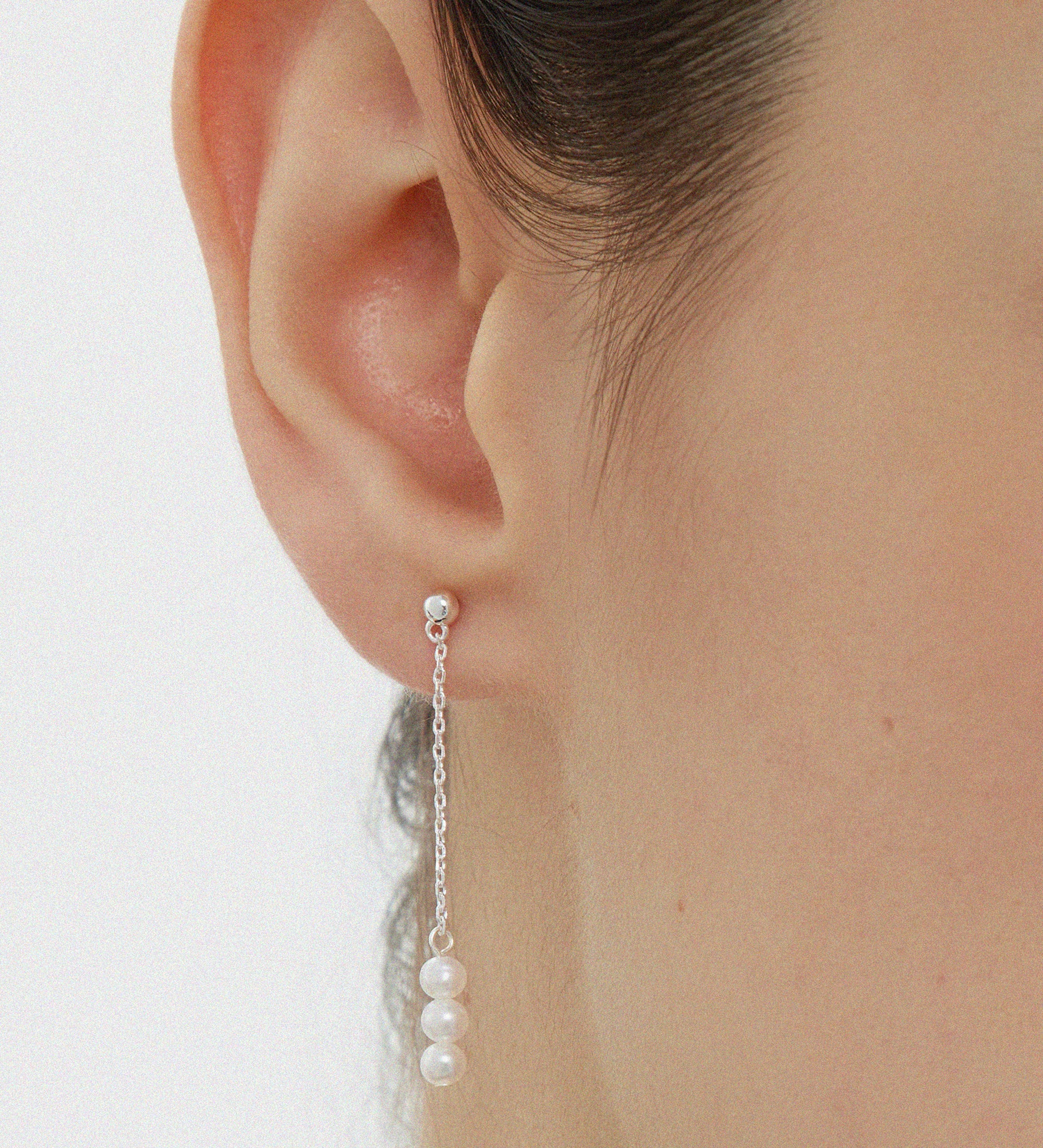 Close-up of a womans ear wearing the Eden Trio Pearl Drop Earrings in Silver, featuring a small stud and three white pearls dangling vertically from a thin silver chain. These elegant earrings stand out against a plain, neutral background.