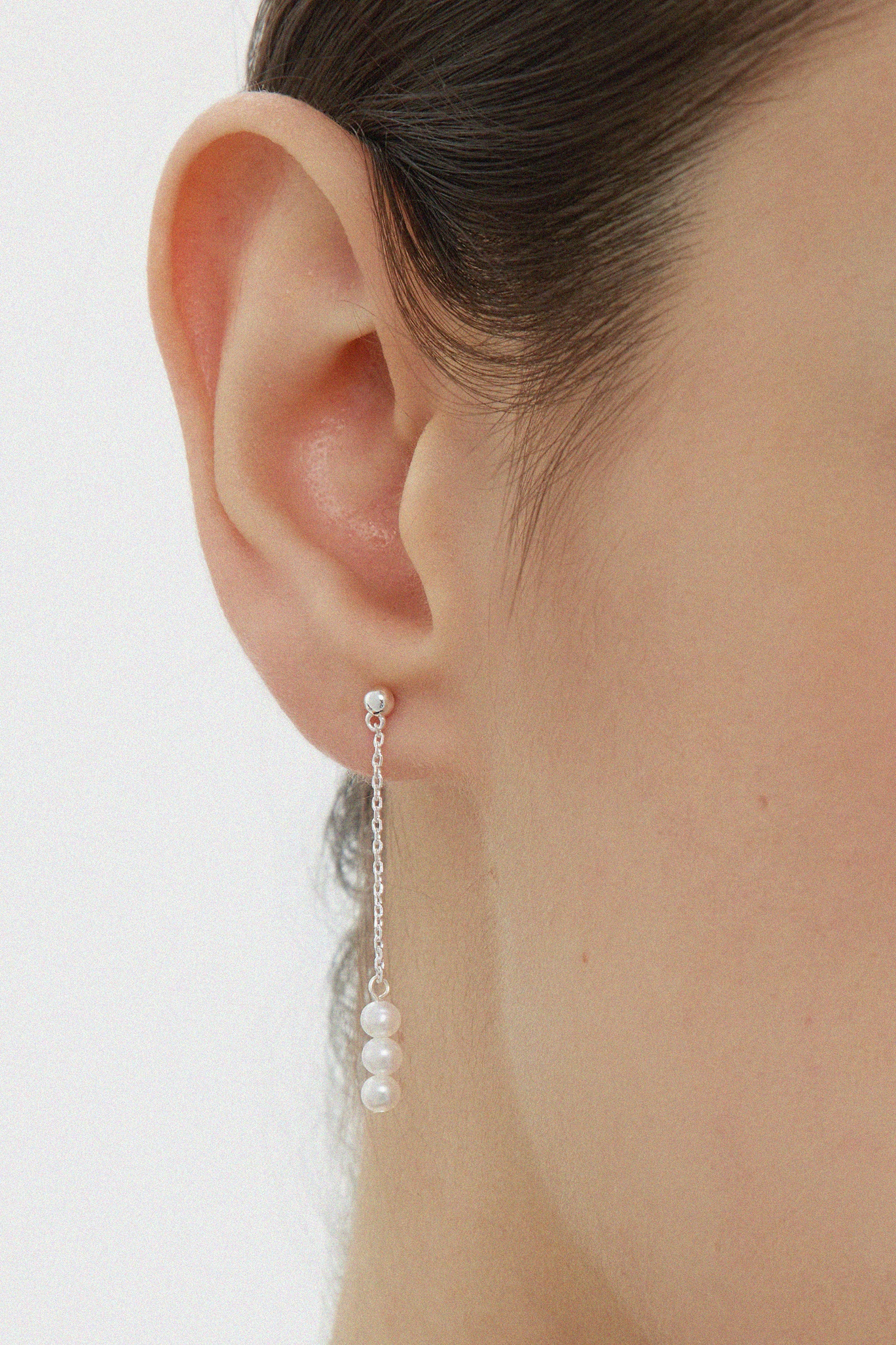 Close-up of a womans ear wearing the Eden Trio Pearl Drop Earrings in Silver, featuring a small stud and three white pearls dangling vertically from a thin silver chain. These elegant earrings stand out against a plain, neutral background.