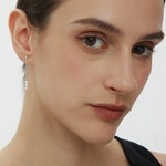 A close-up of a woman with fair skin and brown hair pulled back, wearing a black top and the Eden Trio Pearl Drop Earrings in Silver, looking slightly to the side with a neutral expression.