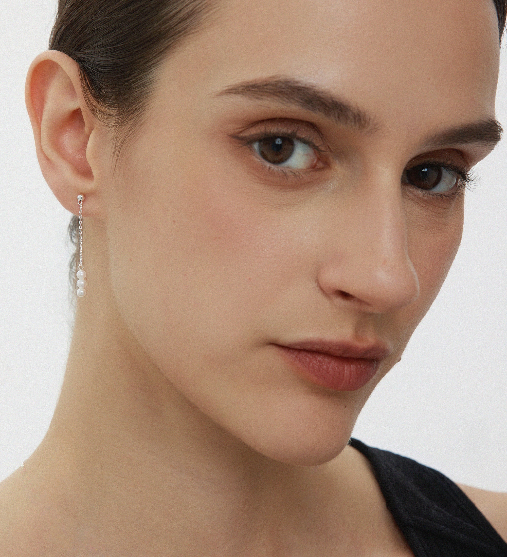 A close-up of a woman with fair skin and brown hair pulled back, wearing a black top and the Eden Trio Pearl Drop Earrings in Silver, looking slightly to the side with a neutral expression.