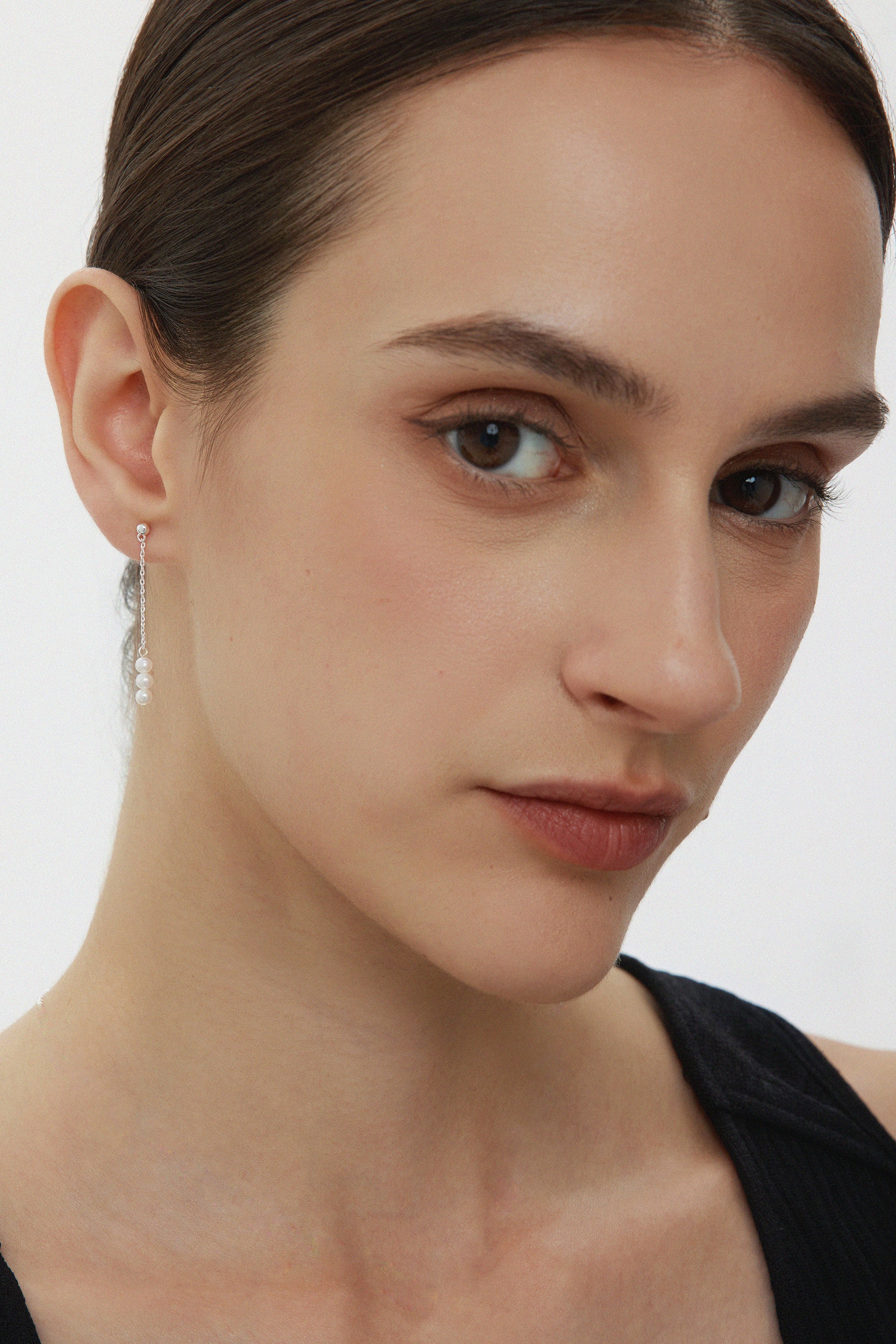 A close-up of a woman with fair skin and brown hair pulled back, wearing a black top and the Eden Trio Pearl Drop Earrings in Silver, looking slightly to the side with a neutral expression.