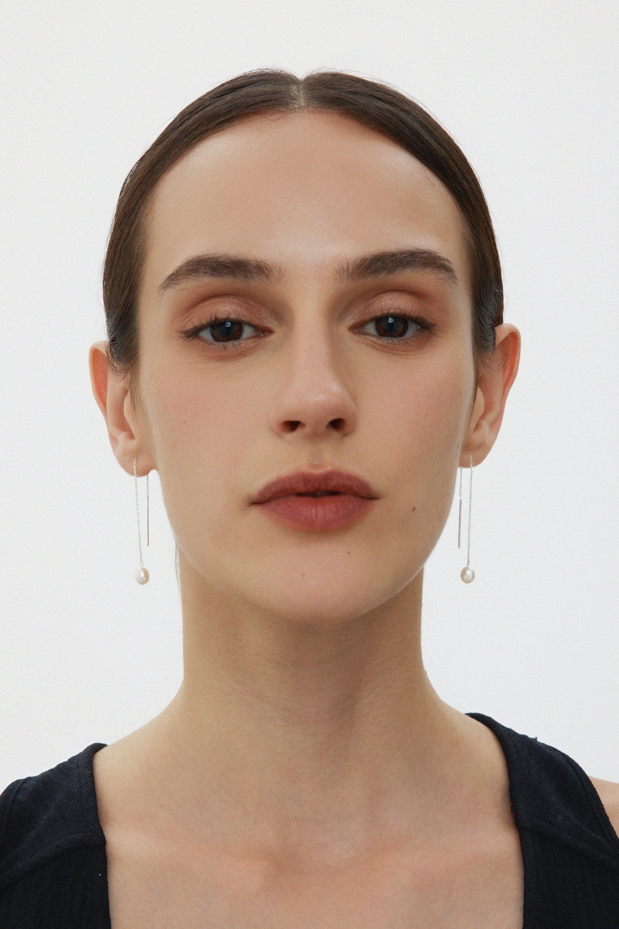 A woman with straight dark hair pulled back wears Eira Pearl Drop Threader Earrings in Silver and a sleeveless black top, facing forward against a plain white background.