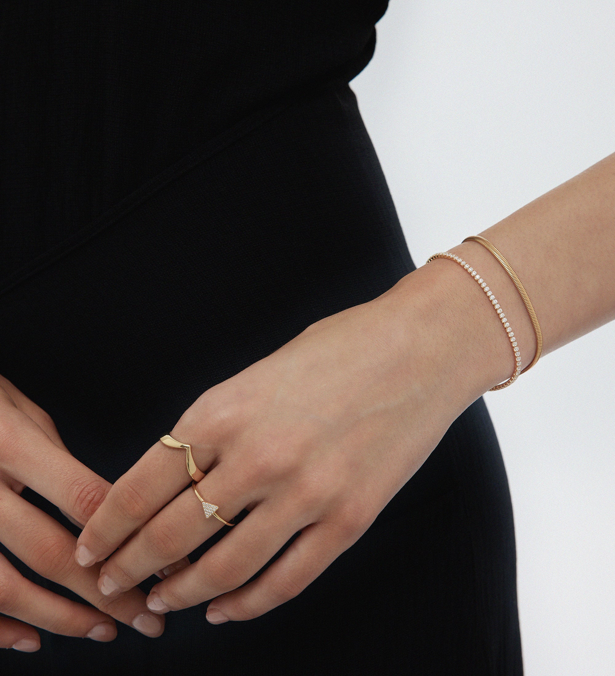 A person in a black dress shows their left hand adorned with two gold rings and a stylish bracelet stack, featuring the Estelle Herringbone Chain Bracelet in Gold alongside delicate pieces, with fingers gently resting on their right hand.