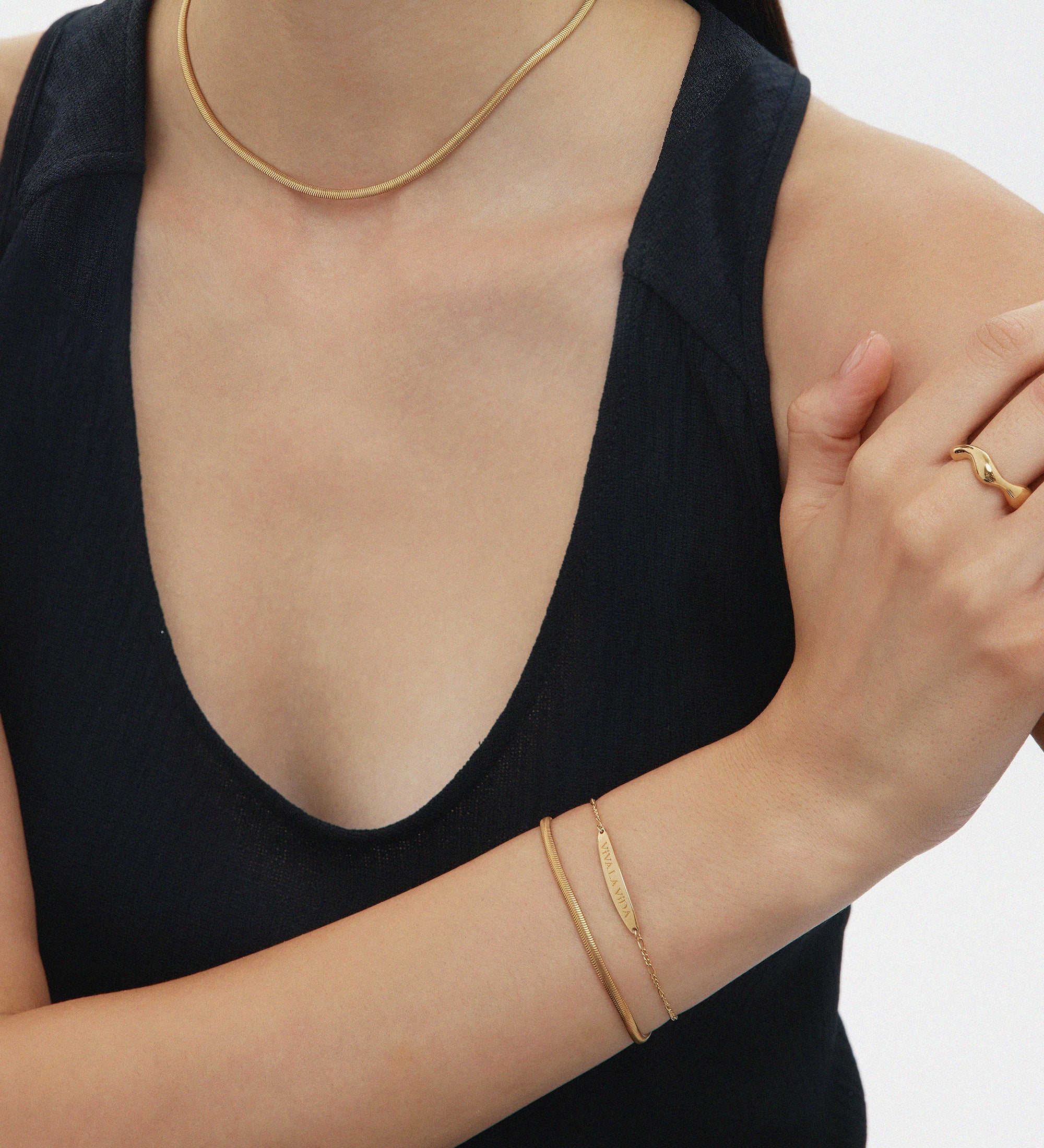 A woman in a sleeveless black top displays gold jewelry: a 3mm chain necklace, the Estelle Herringbone Chain Bracelet in Gold stacked with others, and a ring; her arms are gently crossed, her face partially out of frame.