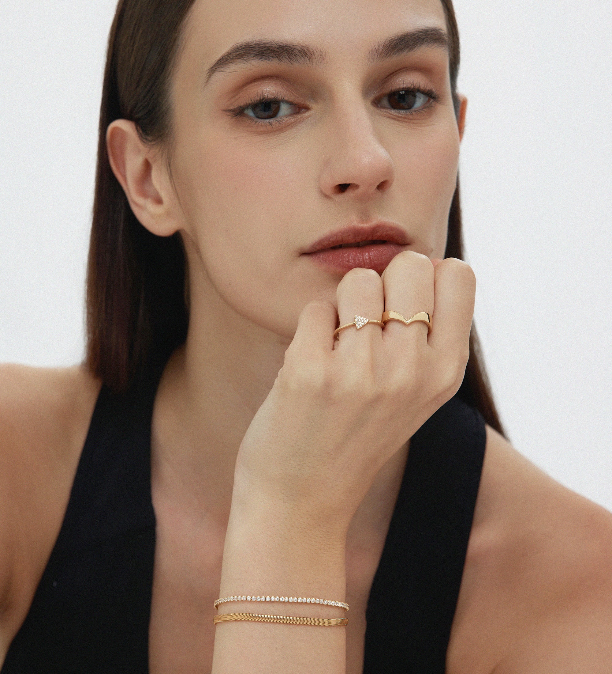 A woman with straight brown hair and light skin wears a black sleeveless top, gold rings, and the Estelle Herringbone Chain Bracelet in Gold as part of her chic bracelet stack, resting her chin on her hand and gazing confidently at the camera.