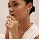 A woman with her hair pulled back wears the Esther Square Hoop Stud Earrings in Silver and a matching ring, resting her hand near her chin. She is dressed in a light sleeveless striped top, gazing thoughtfully into the distance.