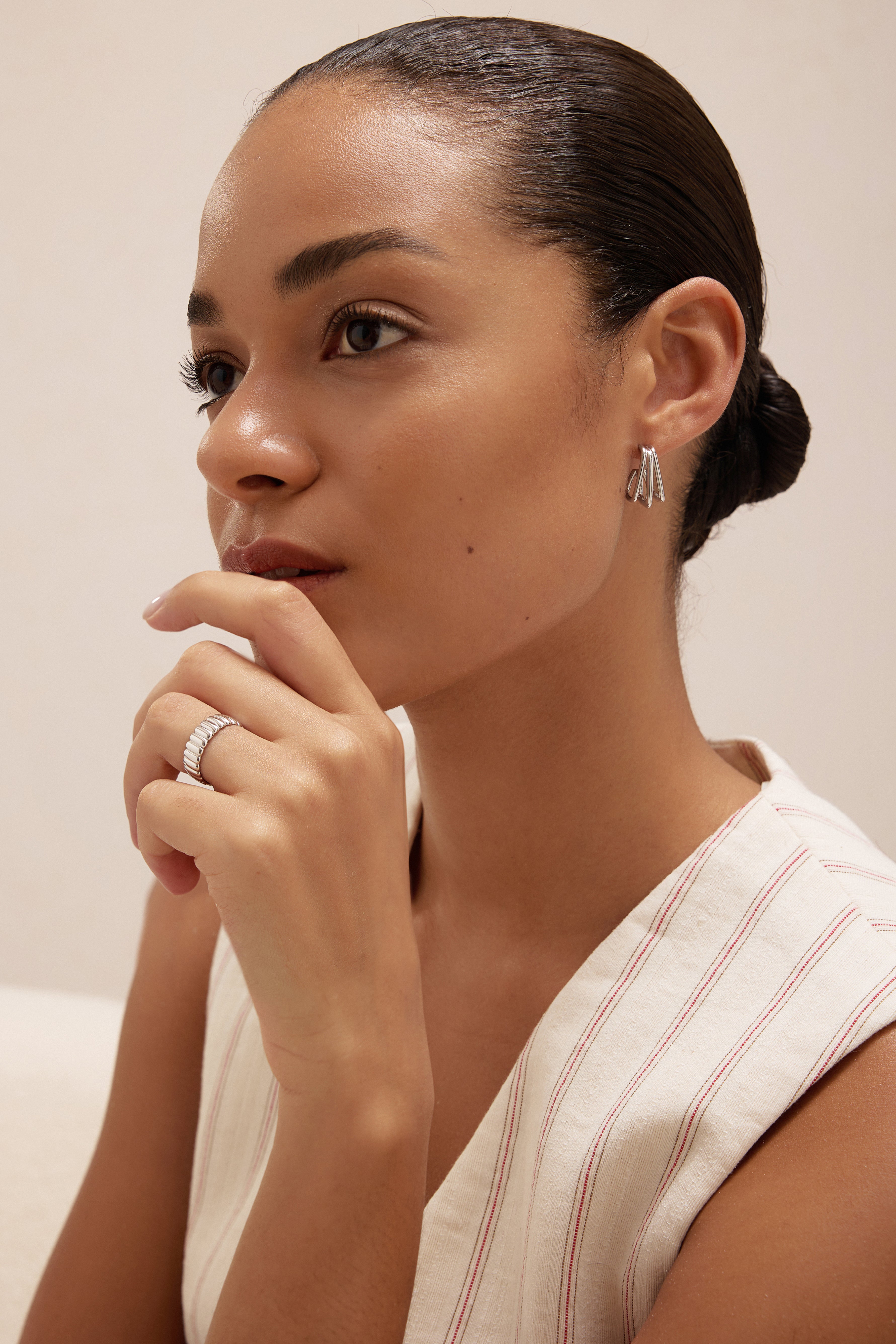 A woman with her hair pulled back wears the Esther Square Hoop Stud Earrings in Silver and a matching ring, resting her hand near her chin. She is dressed in a light sleeveless striped top, gazing thoughtfully into the distance.