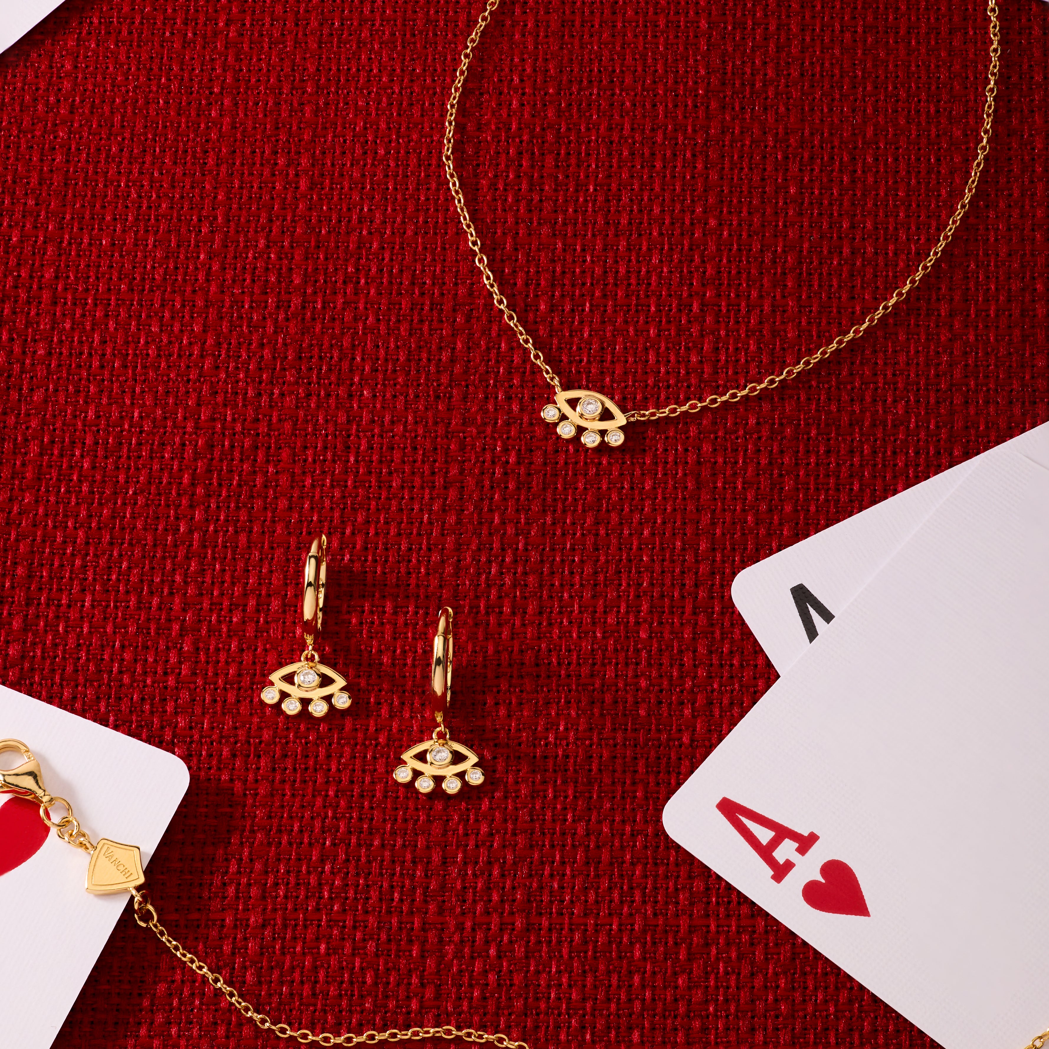 Silver Defender Evil Eye Necklace arranged with matching gold jewelry—bracelet, earrings, and charms—on a red textured surface, surrounded by playing cards like the ace of hearts and king.