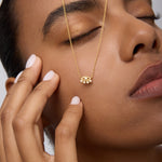 A woman with manicured nails and closed eyes gently touches her face, wearing the Defender Evil Eye Necklace in Gold. The 18k gold-plated pendant rests on her cheek against a neutral background.