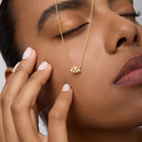 A woman with manicured nails and closed eyes gently touches her face, wearing the Defender Evil Eye Necklace in Gold. The 18k gold-plated pendant rests on her cheek against a neutral background.