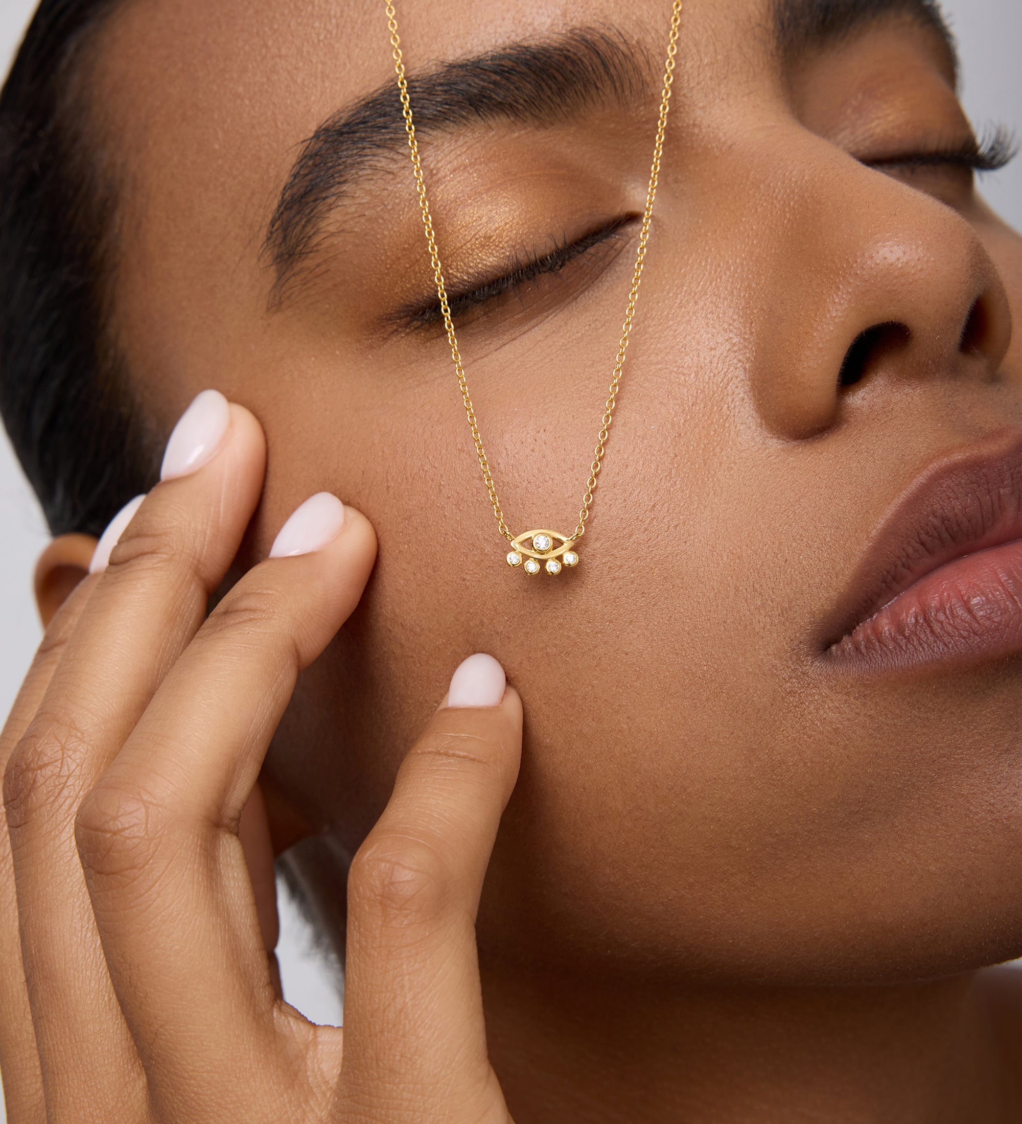 A woman with manicured nails and closed eyes gently touches her face, wearing the Defender Evil Eye Necklace in Gold. The 18k gold-plated pendant rests on her cheek against a neutral background.