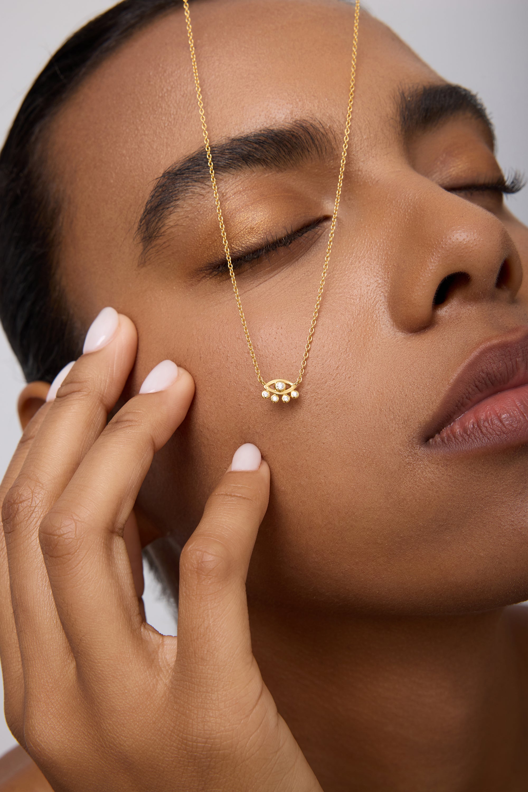 A woman with manicured nails and closed eyes gently touches her face, wearing the Defender Evil Eye Necklace in Gold. The 18k gold-plated pendant rests on her cheek against a neutral background.