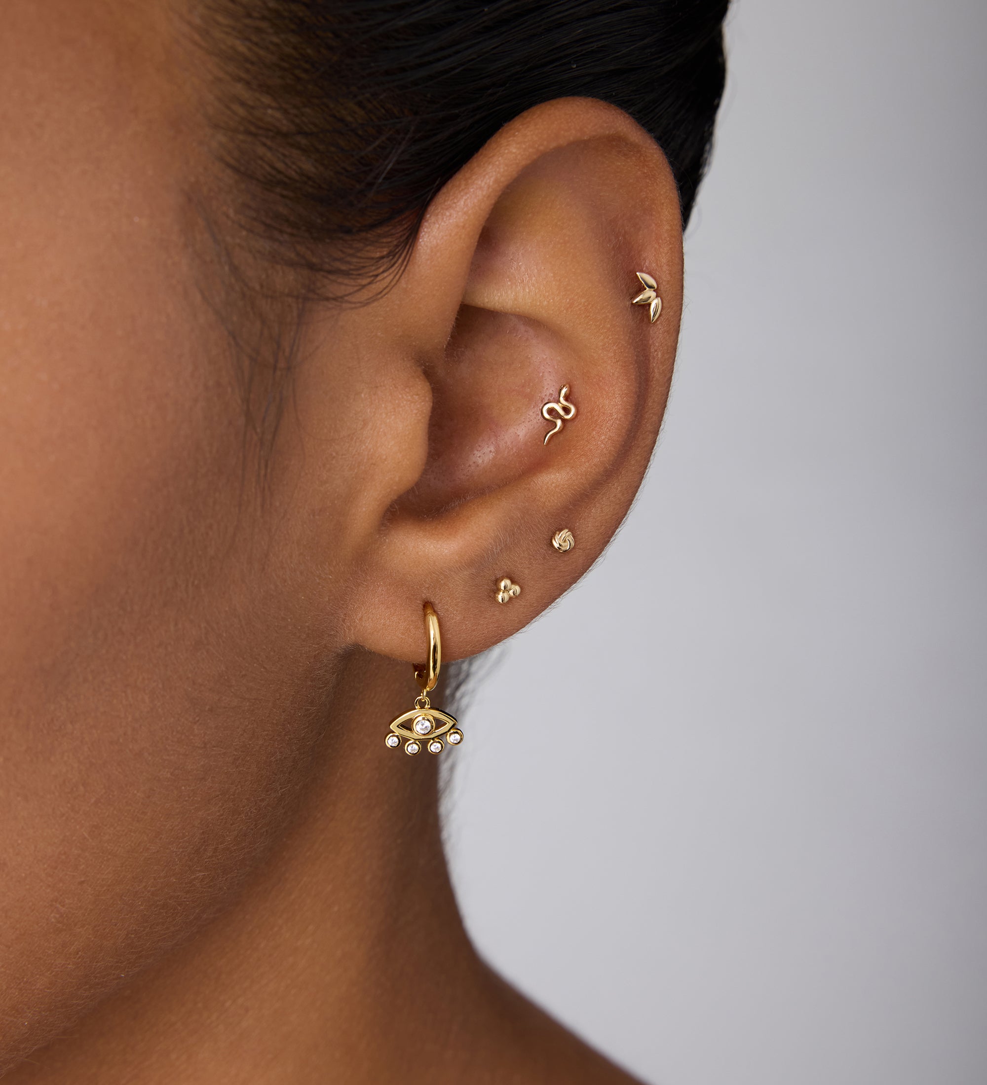 Close-up of an ear adorned with multiple gold earrings, featuring Defender Evil Eye Hoops in Gold with pearl details on the lobe and small protective jewelry along the cartilage.
