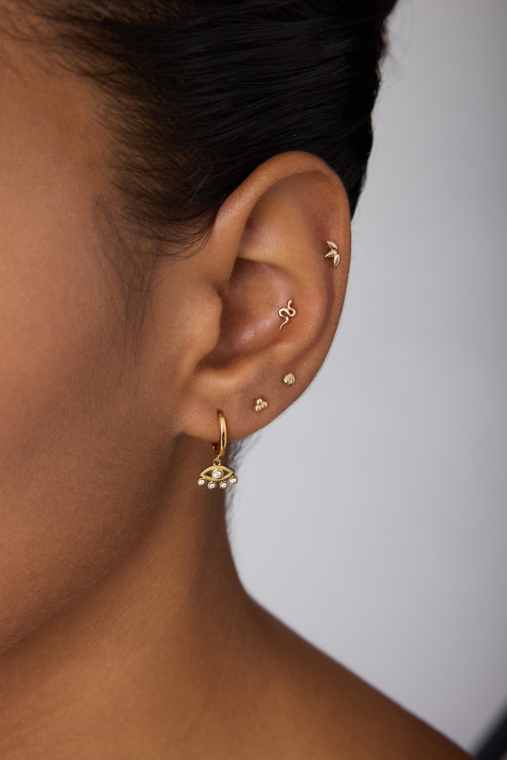 Close-up of an ear adorned with multiple gold earrings, featuring Defender Evil Eye Hoops in Gold with pearl details on the lobe and small protective jewelry along the cartilage.