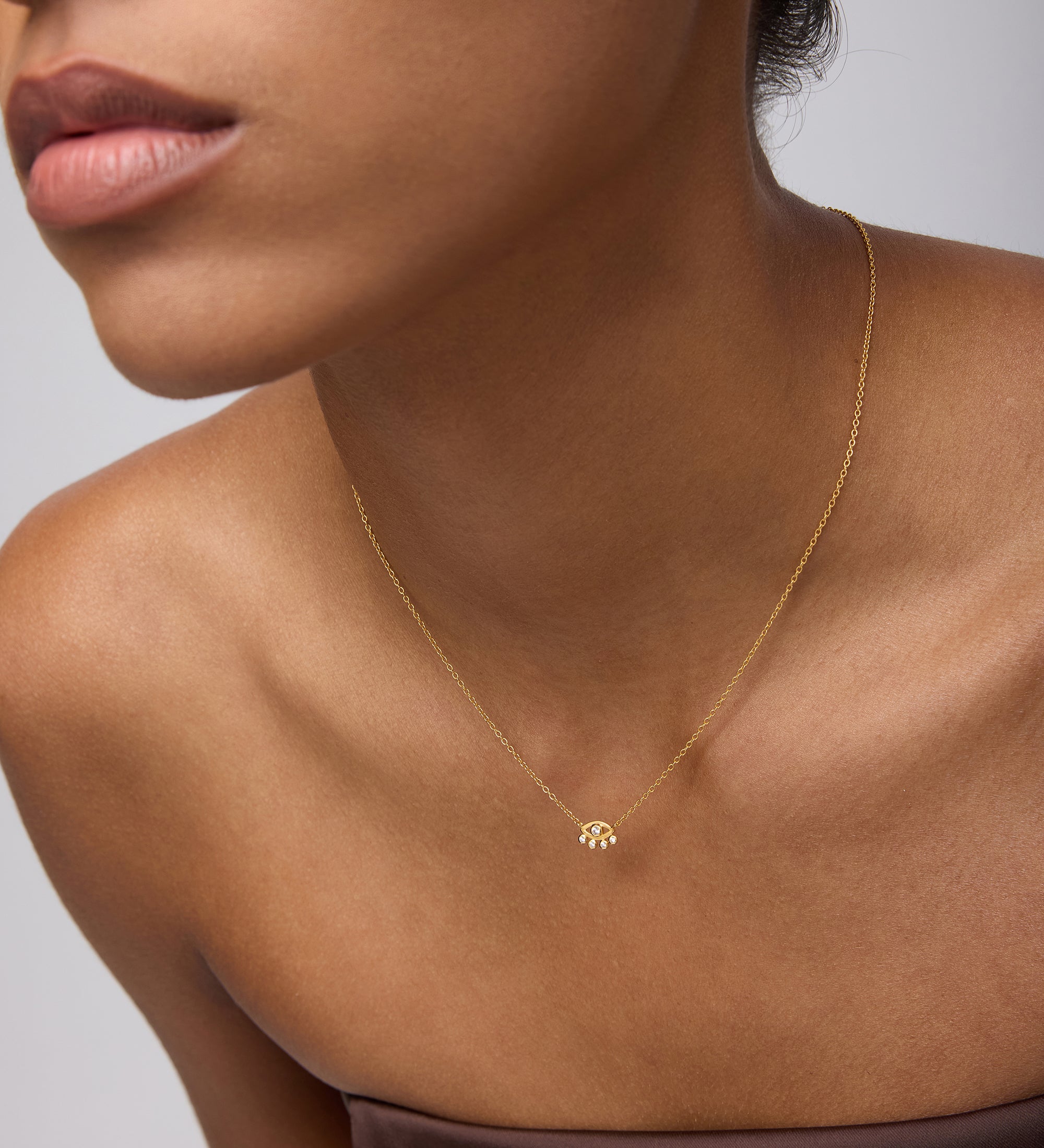 A woman in a strapless brown top showcases the Defender Evil Eye Necklace in Gold, 18k gold plated with a small cubic zirconia pendant, against a neutral background. Her face is partially visible, emphasizing her lips and jawline.