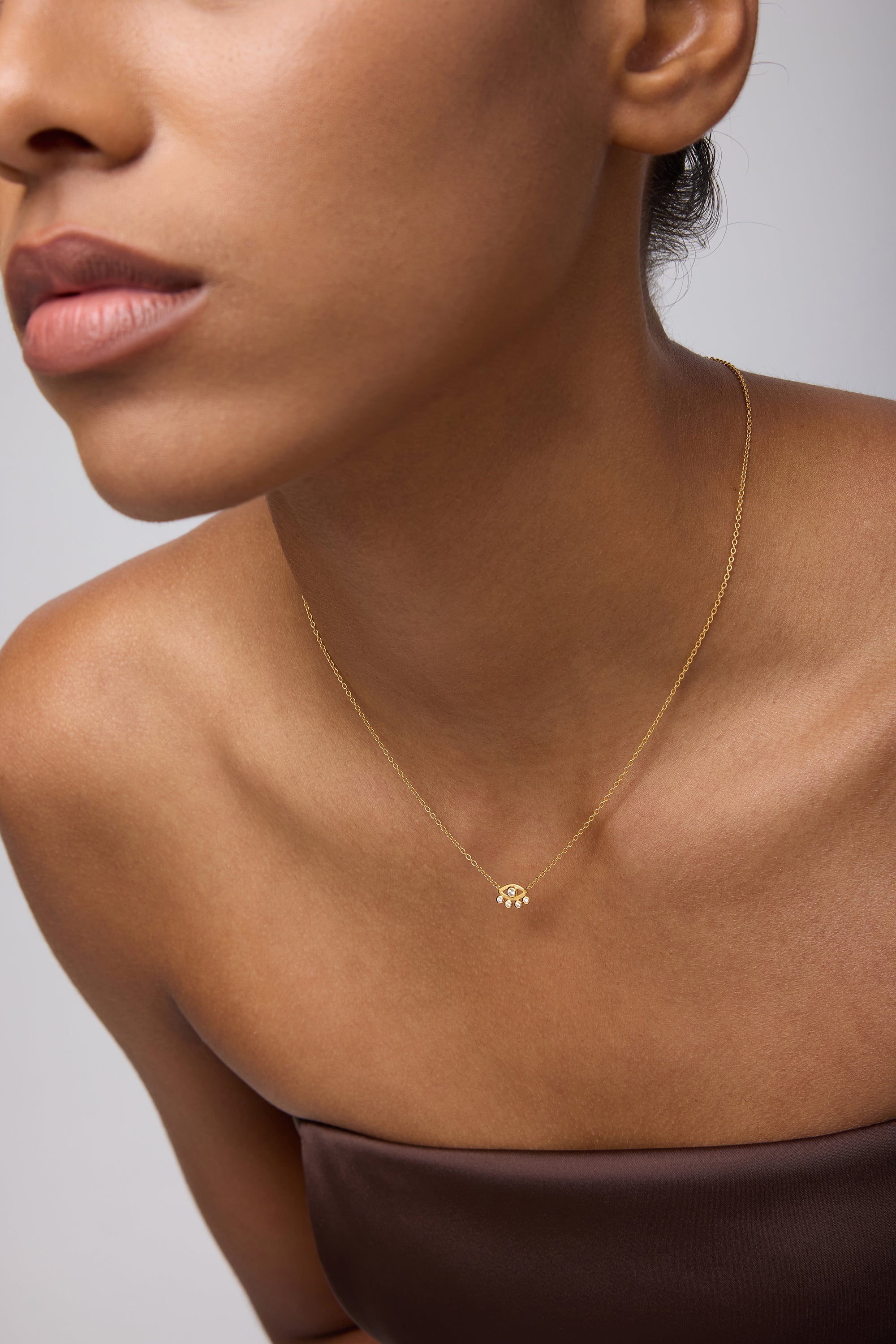A woman in a strapless brown top showcases the Defender Evil Eye Necklace in Gold, 18k gold plated with a small cubic zirconia pendant, against a neutral background. Her face is partially visible, emphasizing her lips and jawline.