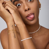 A woman with pulled-back hair playfully sticks out her tongue and widens her eyes, showing off the Defender Evil Eye Bracelet in Gold along with other 18k gold-plated necklaces, earrings, bracelets, and a forearm tattoo.