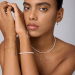 A woman with a neutral expression poses against a light background, highlighting gold hoop earrings, layered gold bracelets, and the Celia Micro Freshwater Pearl Choker Necklace in Silver—her smooth skin and neat brows complete the perfect summer jewelry style.