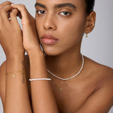 A woman with a neutral expression poses against a light background, highlighting gold hoop earrings, layered gold bracelets, and the Celia Micro Freshwater Pearl Choker Necklace in Silver—her smooth skin and neat brows complete the perfect summer jewelry style.