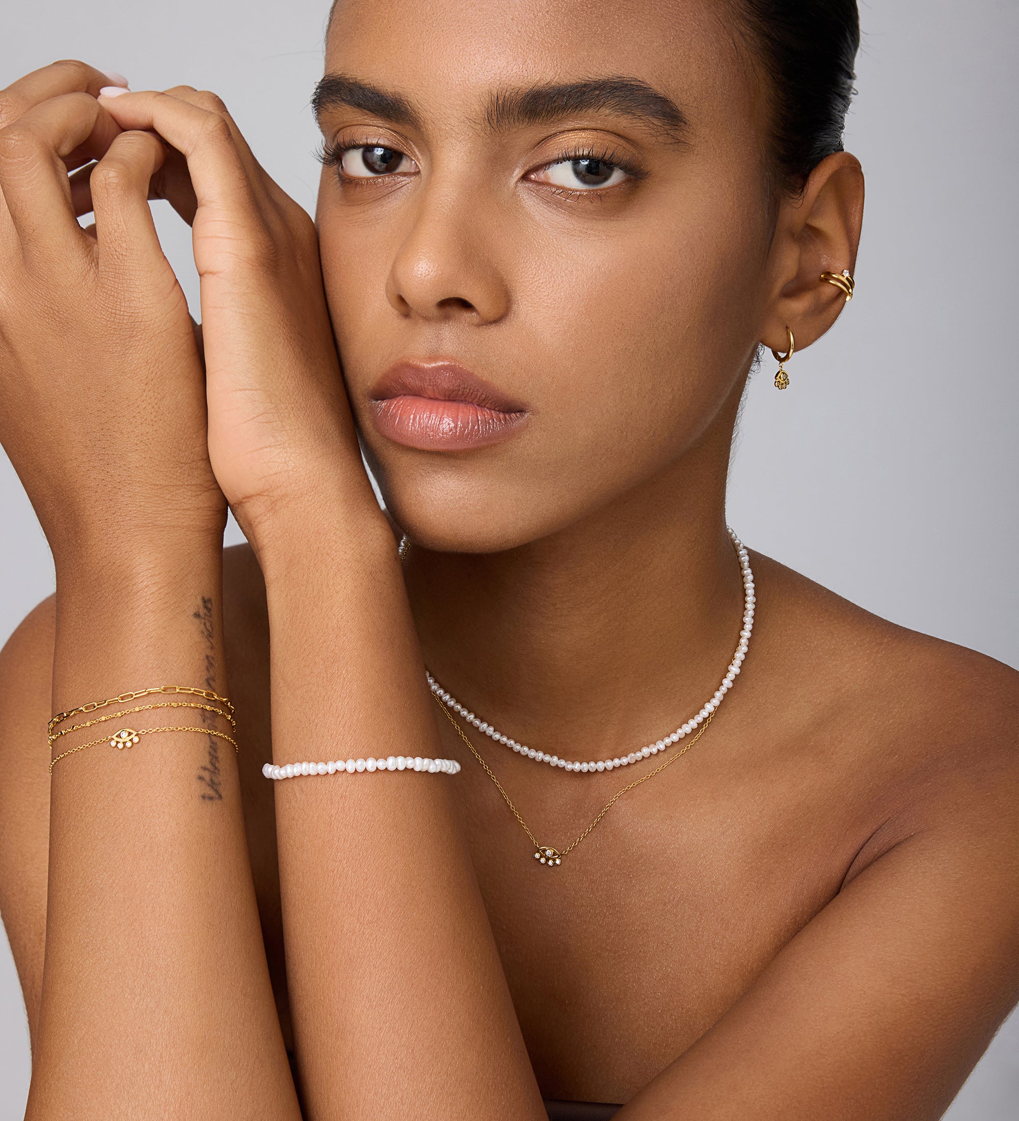 A woman with a neutral expression poses against a light background, highlighting gold hoop earrings, layered gold bracelets, and the Celia Micro Freshwater Pearl Choker Necklace in Silver—her smooth skin and neat brows complete the perfect summer jewelry style.