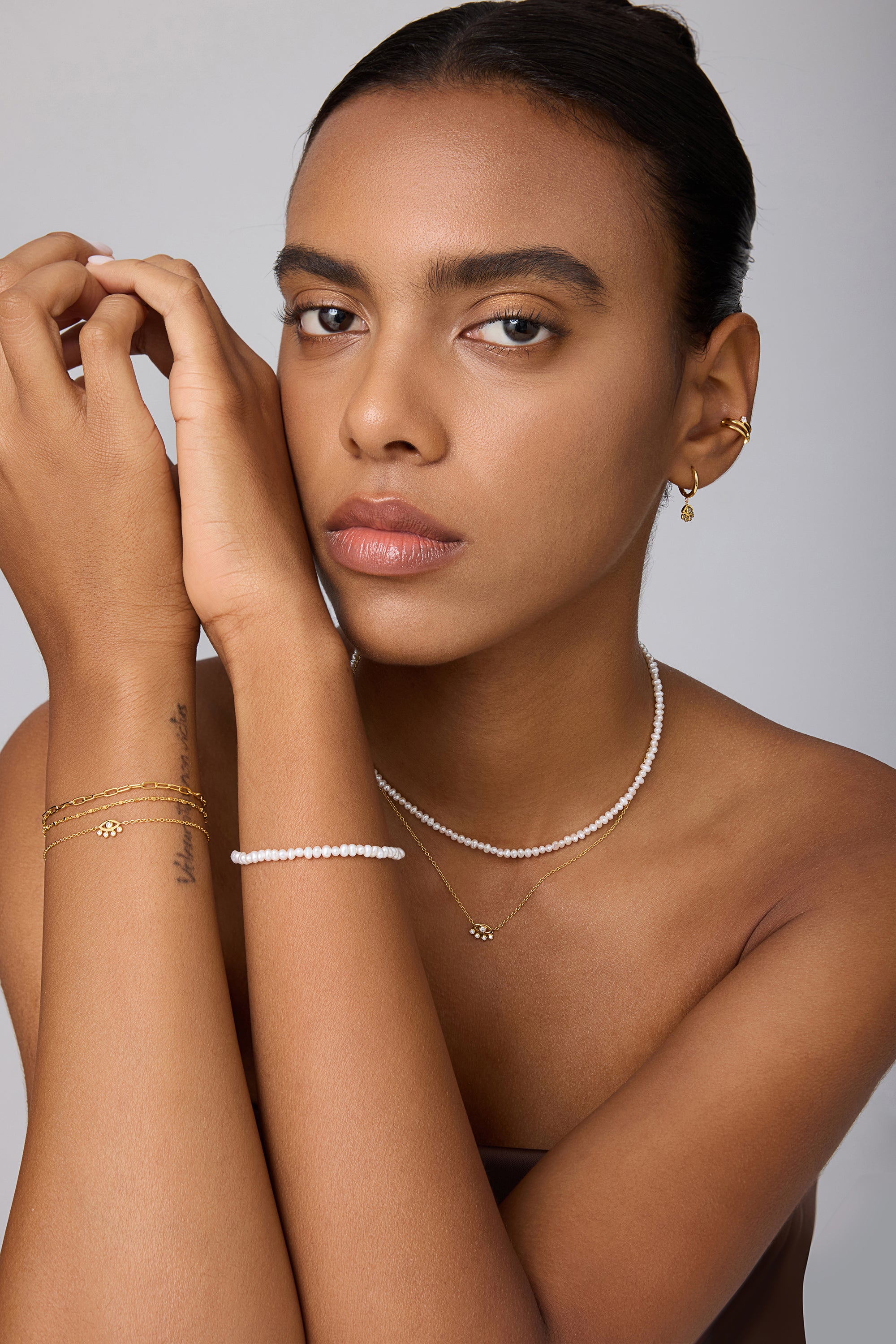 A woman with a neutral expression poses against a light background, highlighting gold hoop earrings, layered gold bracelets, and the Celia Micro Freshwater Pearl Choker Necklace in Silver—her smooth skin and neat brows complete the perfect summer jewelry style.