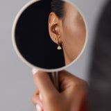 A person holds a round mirror reflecting their ear adorned with gold plated earrings, including studs, a small dangle, and Defender Evil Eye Hoops in Gold, against a plain gray background.