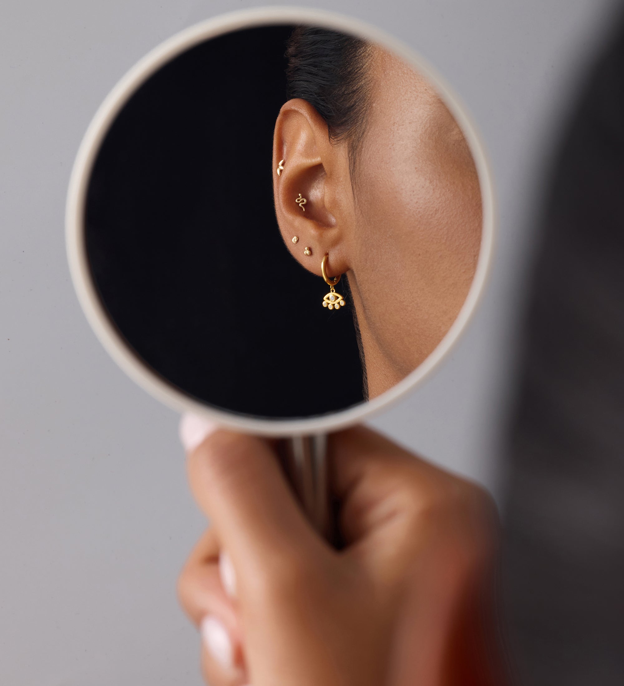 A person holds a round mirror reflecting their ear adorned with gold plated earrings, including studs, a small dangle, and Defender Evil Eye Hoops in Gold, against a plain gray background.
