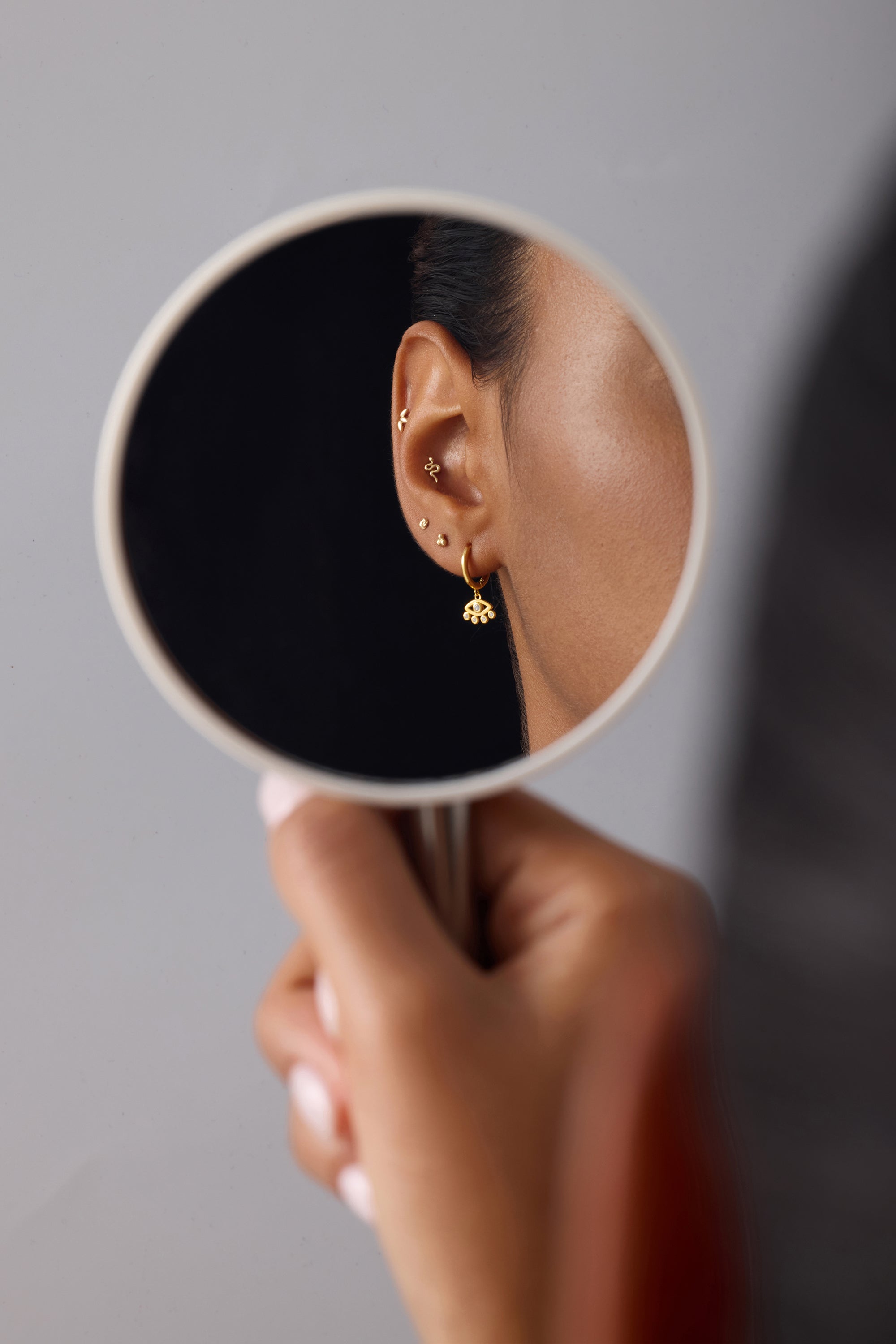 A person holds a round mirror reflecting their ear adorned with gold plated earrings, including studs, a small dangle, and Defender Evil Eye Hoops in Gold, against a plain gray background.