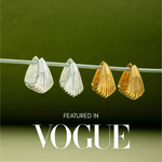 Two pairs of sculptural hoop earrings, including the Juno Ridged Chunky Hoops in Gold, hang on a clear rod against a green background. The text Featured in VOGUE appears at the bottom of the image.
