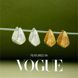 Two pairs of sculptural hoop earrings, including the Juno Ridged Chunky Hoops in Gold, hang on a clear rod against a green background. The text Featured in VOGUE appears at the bottom of the image.