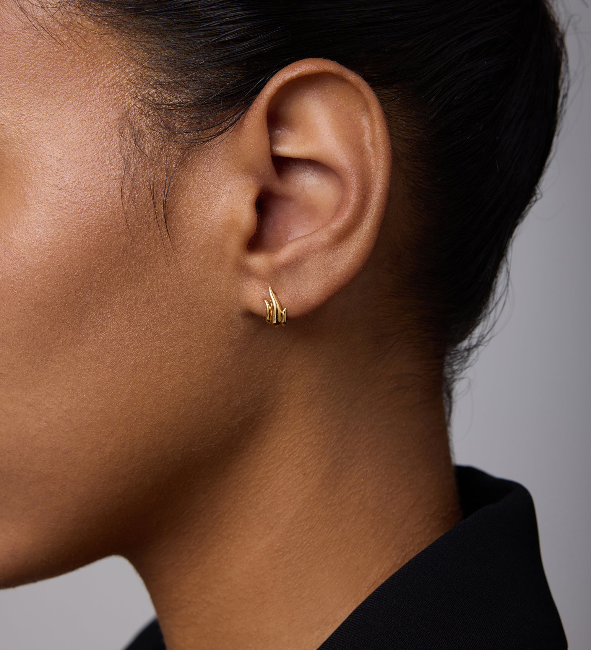 Close-up of a woman wearing Flame Stud Earrings in Gold—small, 18k gold-plated multi-bar studs. Her dark hair is pulled back, she's in a black collared top, and the background is soft and neutral.