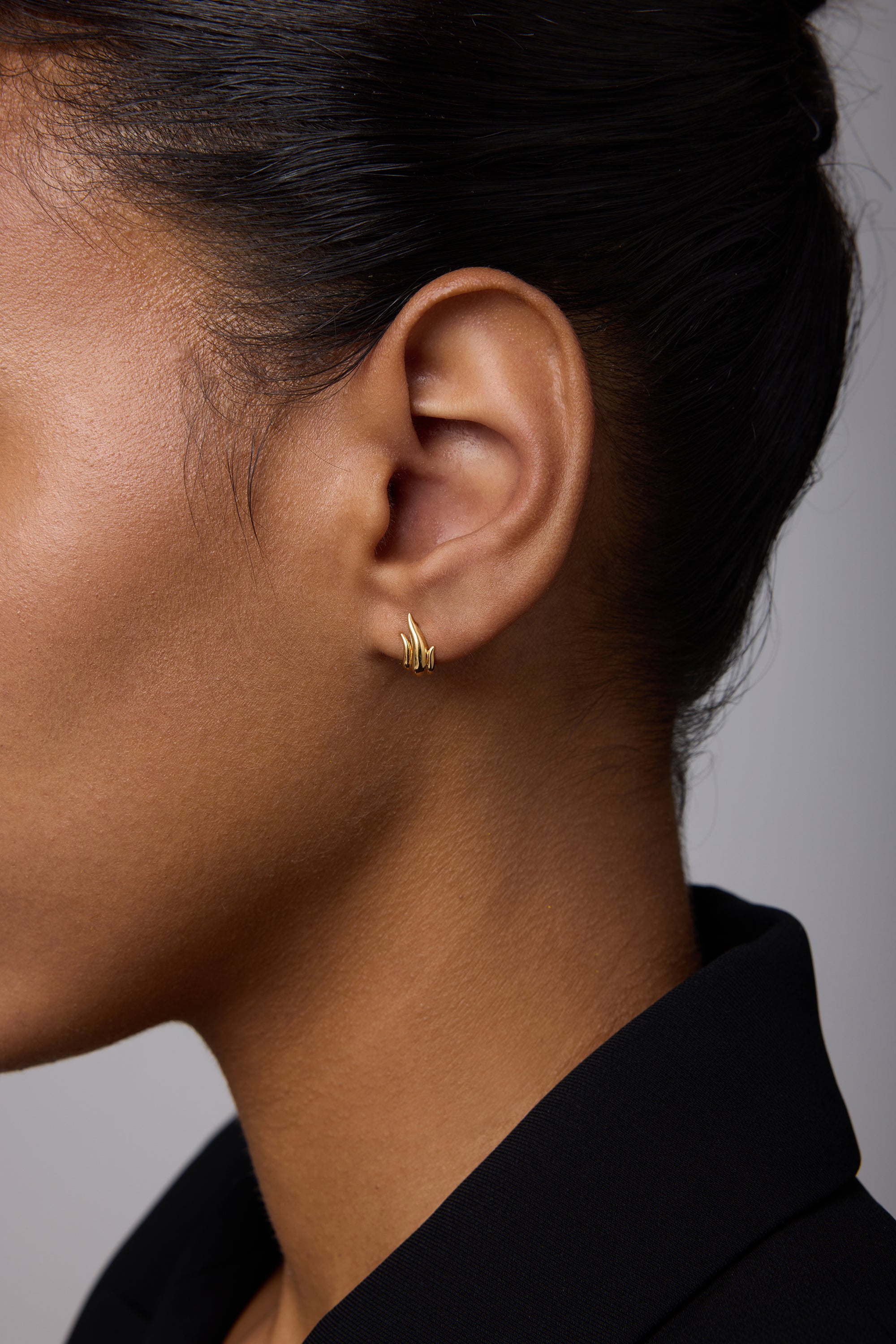 Close-up of a woman wearing Flame Stud Earrings in Gold—small, 18k gold-plated multi-bar studs. Her dark hair is pulled back, she's in a black collared top, and the background is soft and neutral.