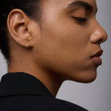 A person with smooth skin and pulled-back hair wears a black blazer, looking to the side. They showcase the Flame Stud Earrings in Gold—small, sculptural earrings with an abstract wave design and an 18k gold-plated finish.