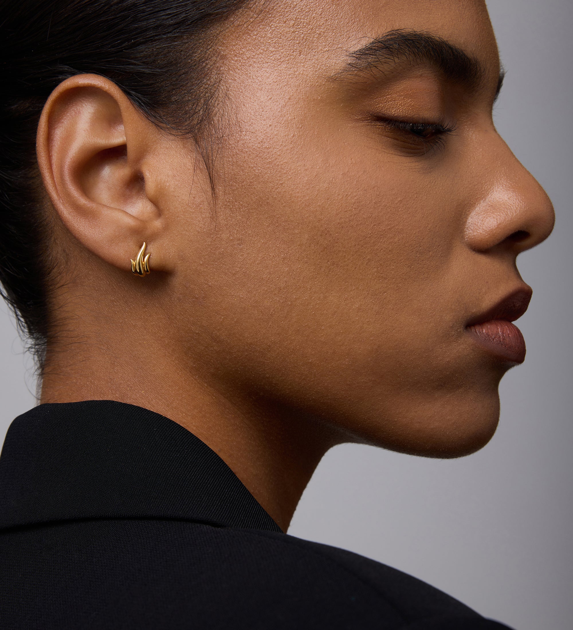 A person with smooth skin and pulled-back hair wears a black blazer, looking to the side. They showcase the Flame Stud Earrings in Gold—small, sculptural earrings with an abstract wave design and an 18k gold-plated finish.