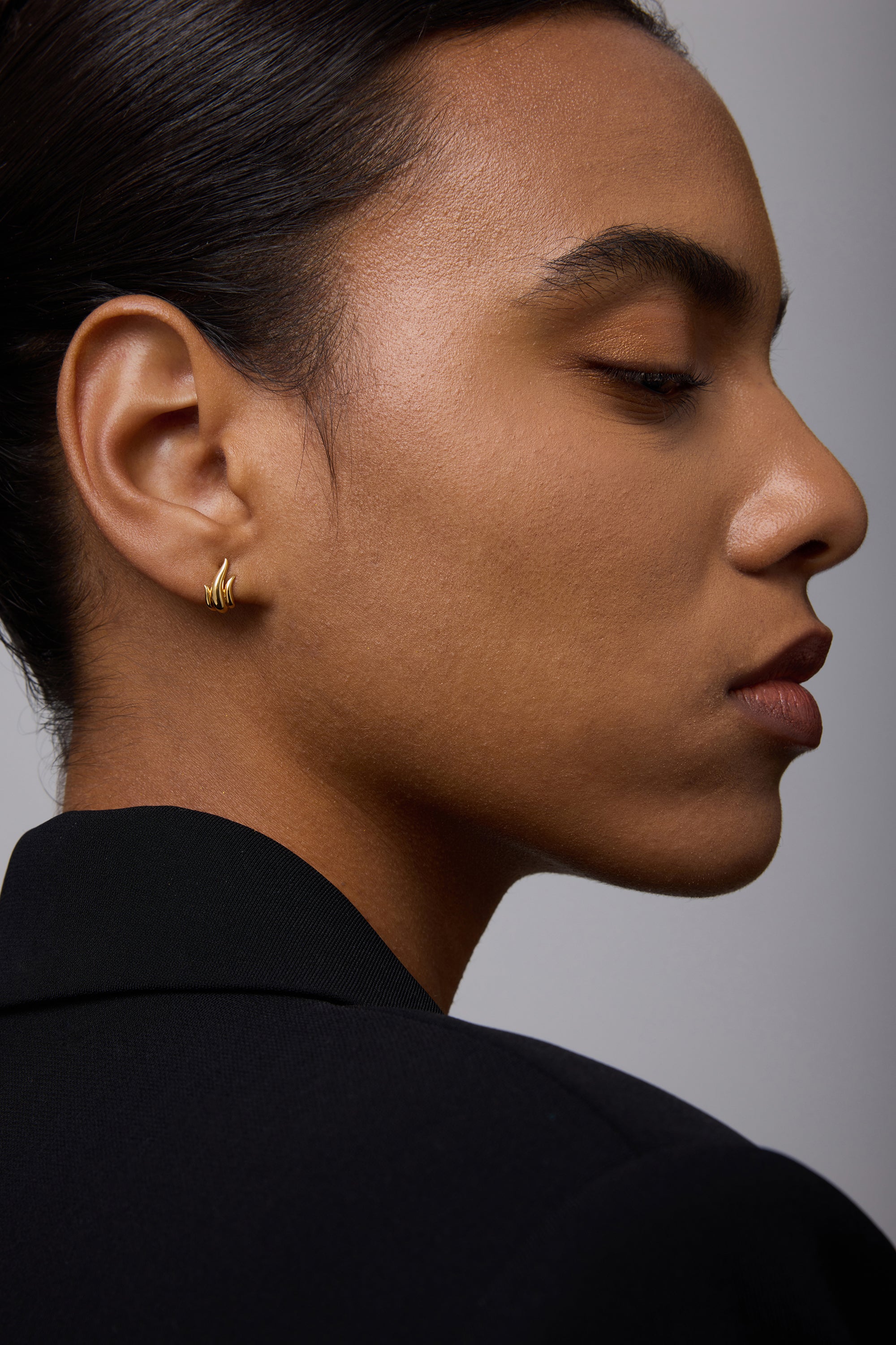 A person with smooth skin and pulled-back hair wears a black blazer, looking to the side. They showcase the Flame Stud Earrings in Gold—small, sculptural earrings with an abstract wave design and an 18k gold-plated finish.