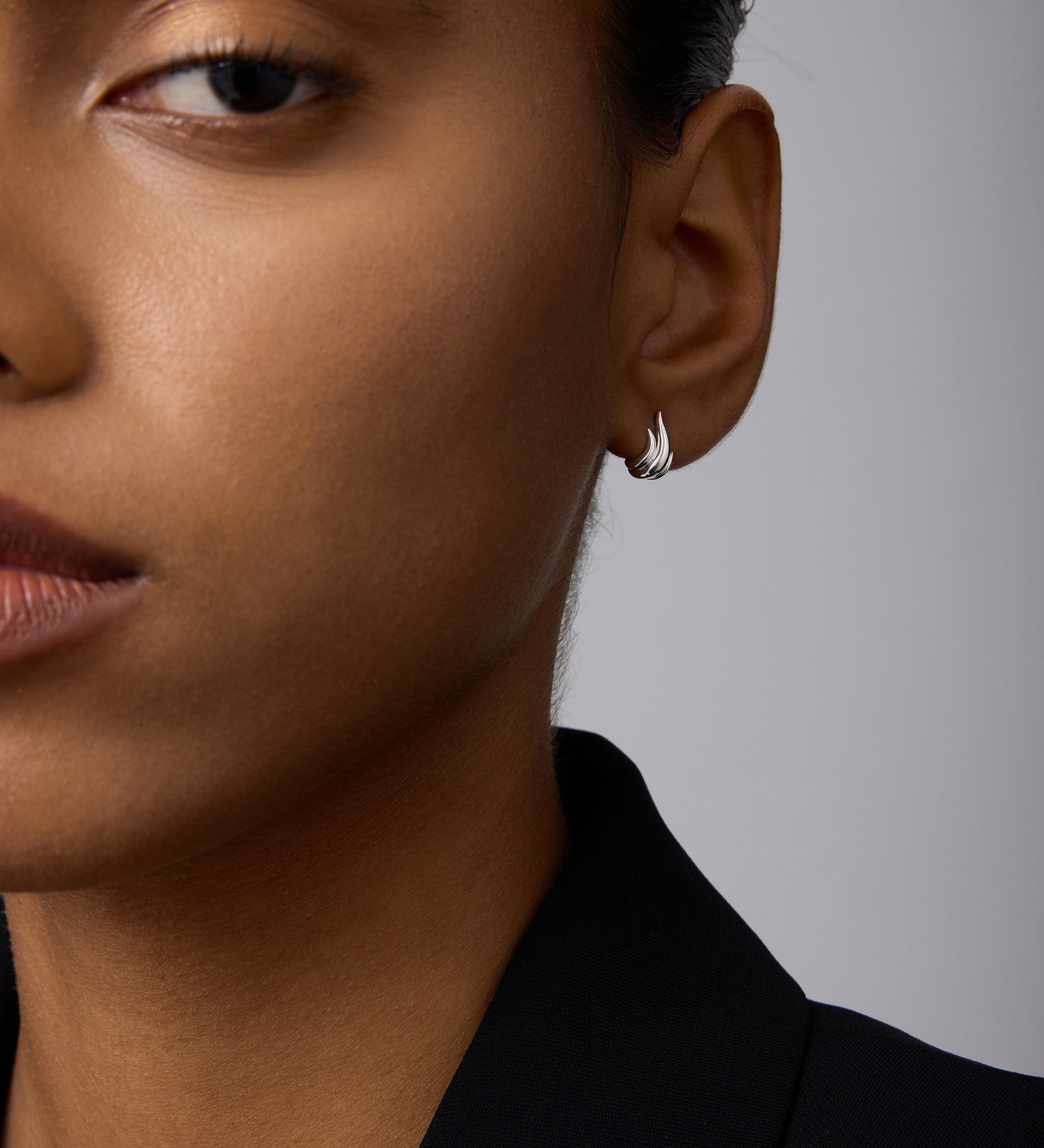 Close-up of a person with medium brown skin wearing Flame Stud Earrings in Silver and a black blazer, hair pulled back, with only the left side of their face visible.