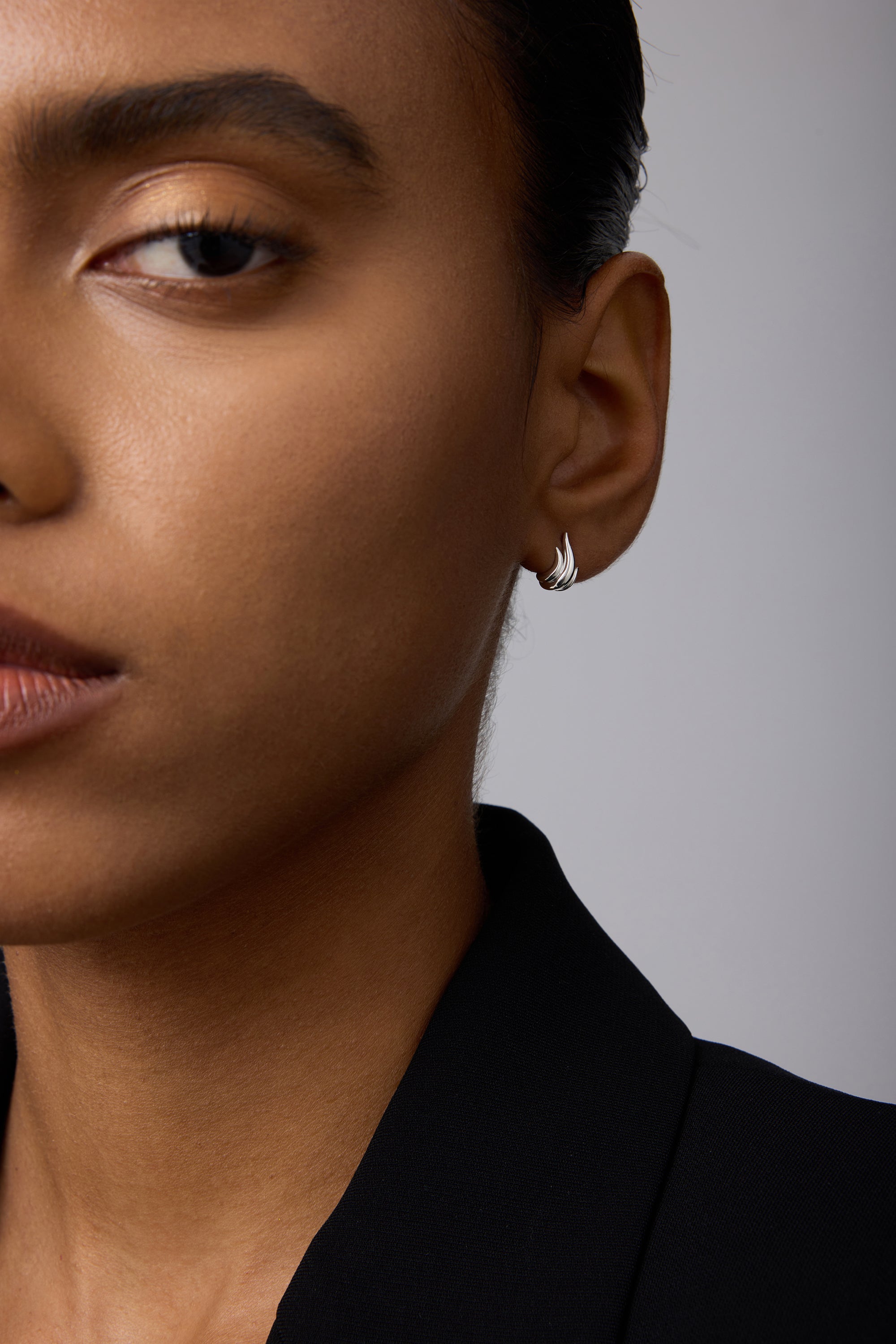 Close-up of a person with medium brown skin wearing Flame Stud Earrings in Silver and a black blazer, hair pulled back, with only the left side of their face visible.