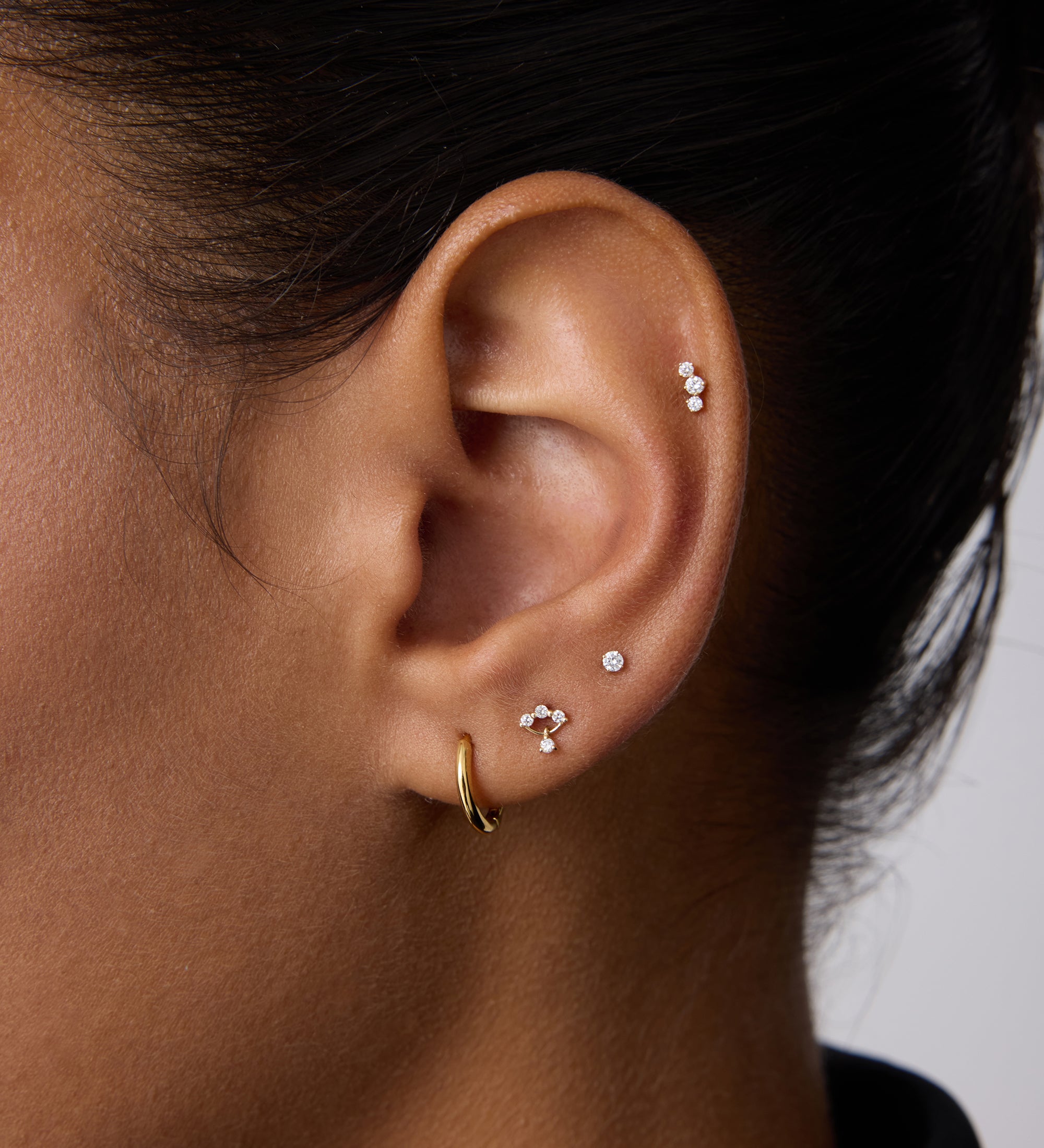 Close-up of a person's ear with multiple piercings, showcasing three Freda Lab-grown Diamond Flat Back Studs in 14K solid yellow gold on the upper and middle ear, plus a gold hoop on the lobe. The person has short dark hair.