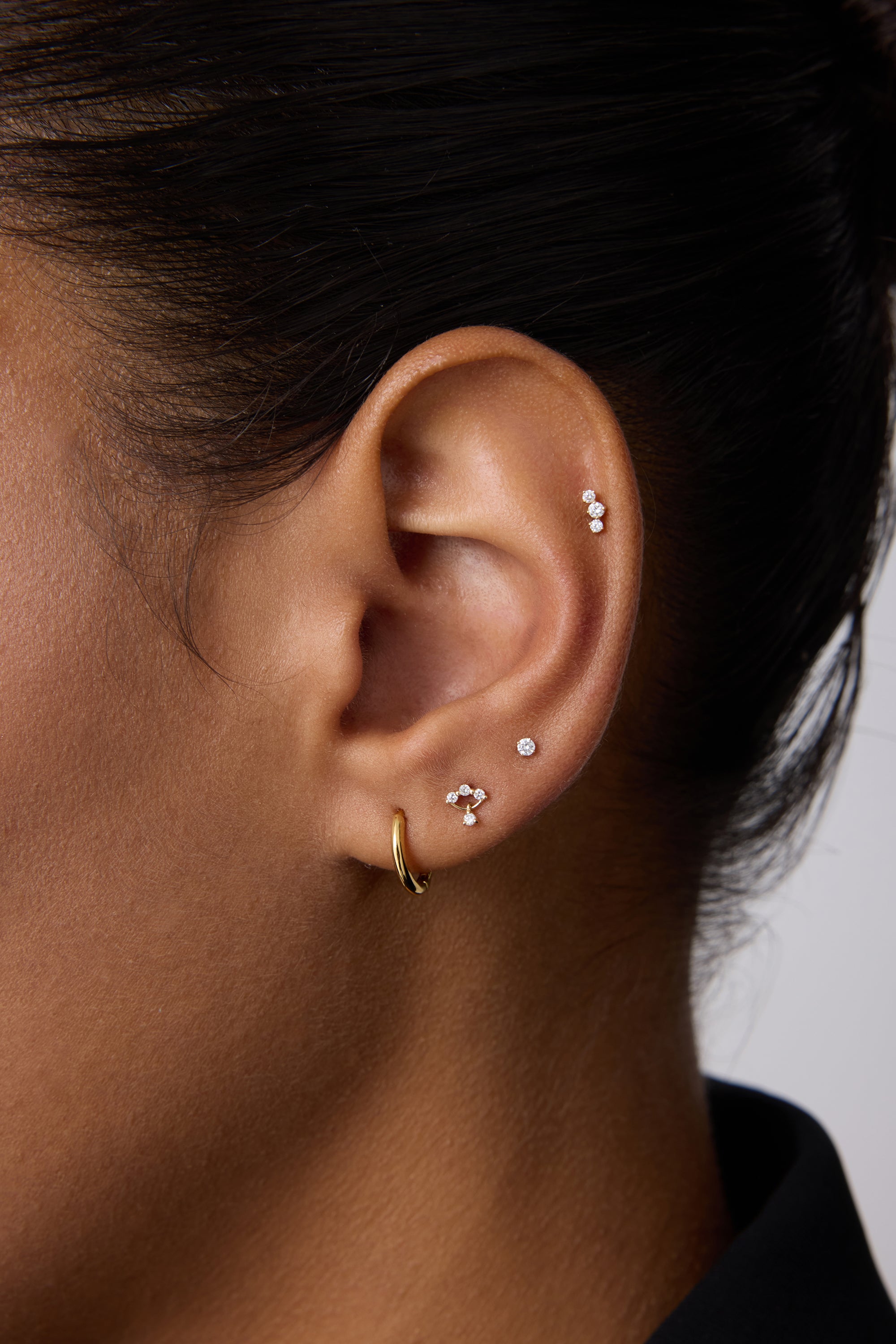 Close-up of a person's ear with multiple piercings, showcasing three Freda Lab-grown Diamond Flat Back Studs in 14K solid yellow gold on the upper and middle ear, plus a gold hoop on the lobe. The person has short dark hair.