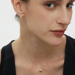 A woman with fair skin and brown hair pulled back wears a black top, a delicate gold necklace, and Georgie Crystal Knife Edge Huggie Hoop Earrings in Gold. She gazes softly to the side with a neutral expression.